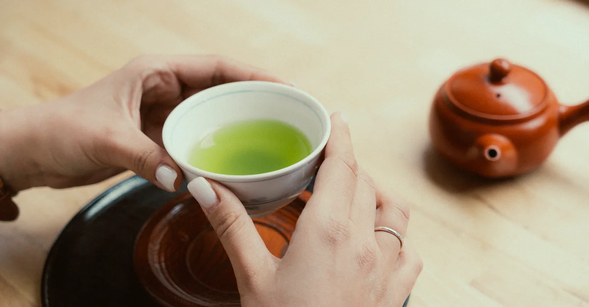 Des mains tenant une tasse de thé vert à côté d'une théière japonaise traditionnelle.