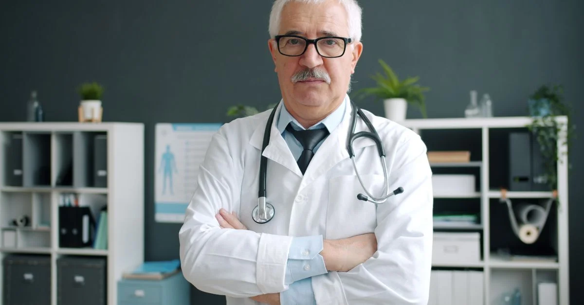 Un médecin, debout avec assurance dans son cabinet médical, stéthoscope à la main, les bras croisés.