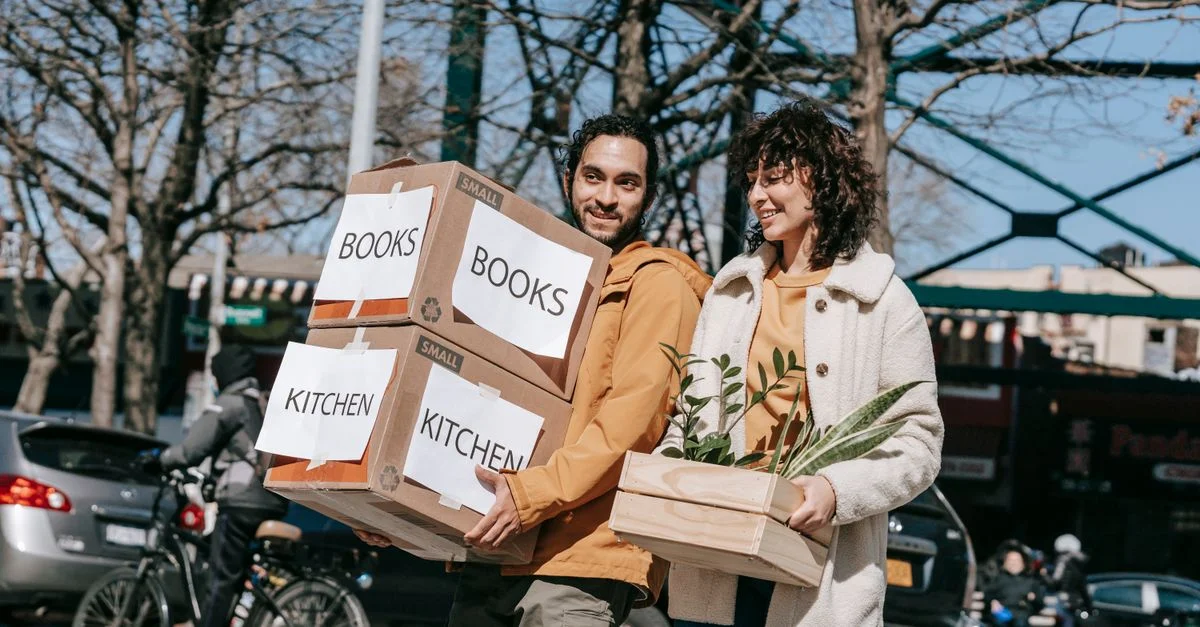 Un couple joyeux portant des cartons étiquetés livres et cuisine dans un cadre urbain, symbolisant un nouveau départ.