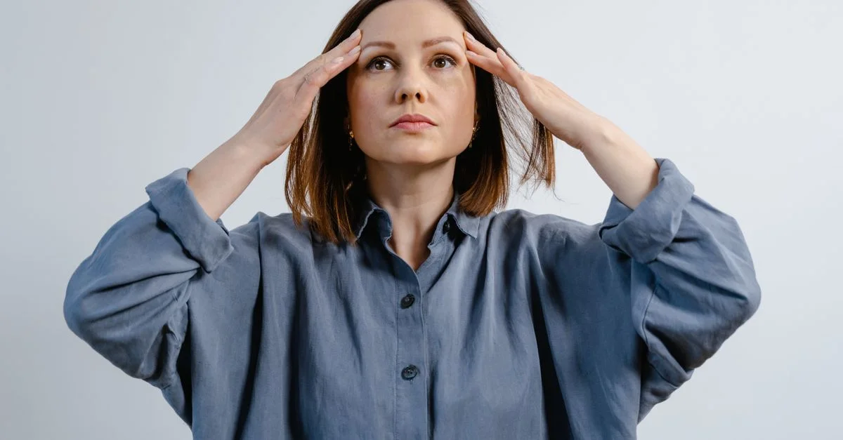 Femme en chemise grise avec les mains sur la tête, regardant pensivement vers le haut à l'intérieur.