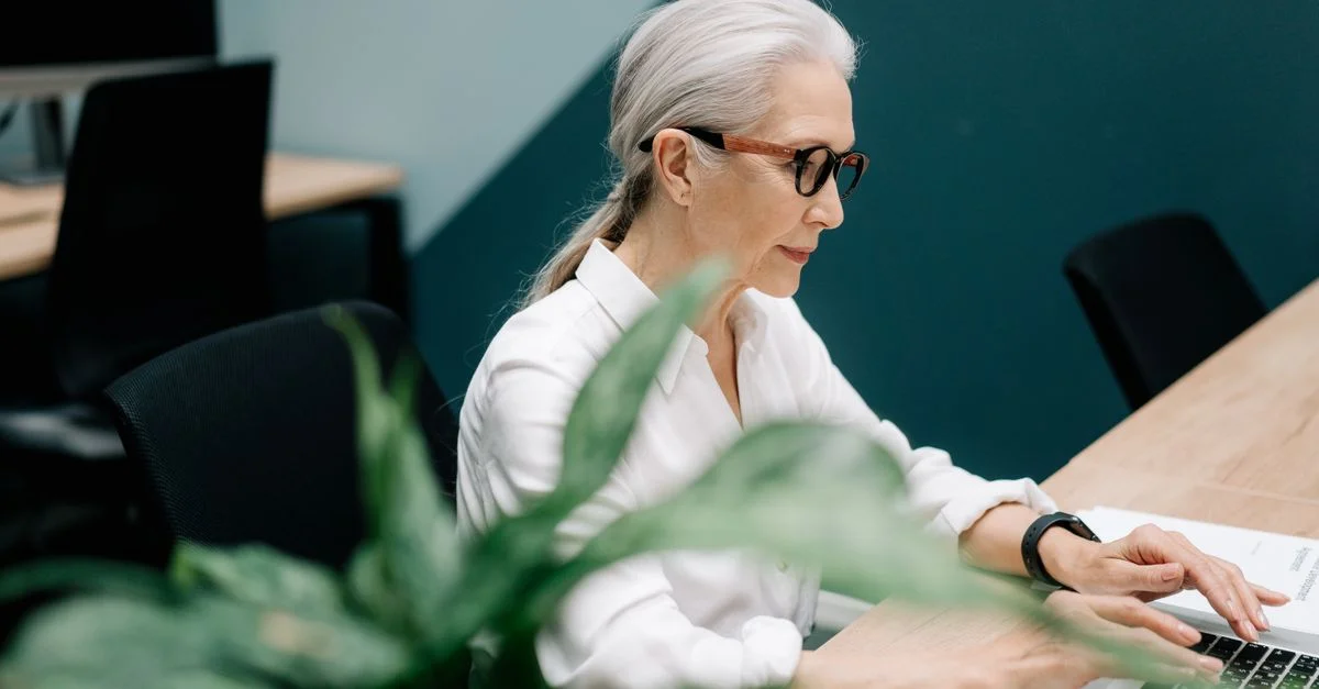 Femme senior confiante portant des lunettes travaillant sur un ordinateur portable dans un bureau moderne.