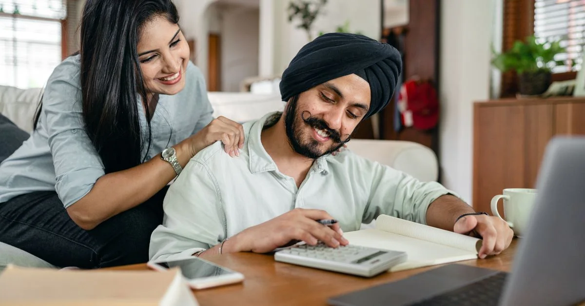 Un couple souriant collabore sur des documents financiers à la maison, utilisant un ordinateur portable et une calculatrice.