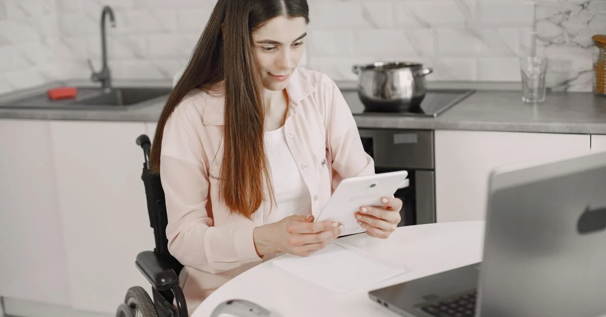 Femme en fauteuil roulant engagée avec une tablette à la table de cuisine, créant un environnement de travail inclusif.