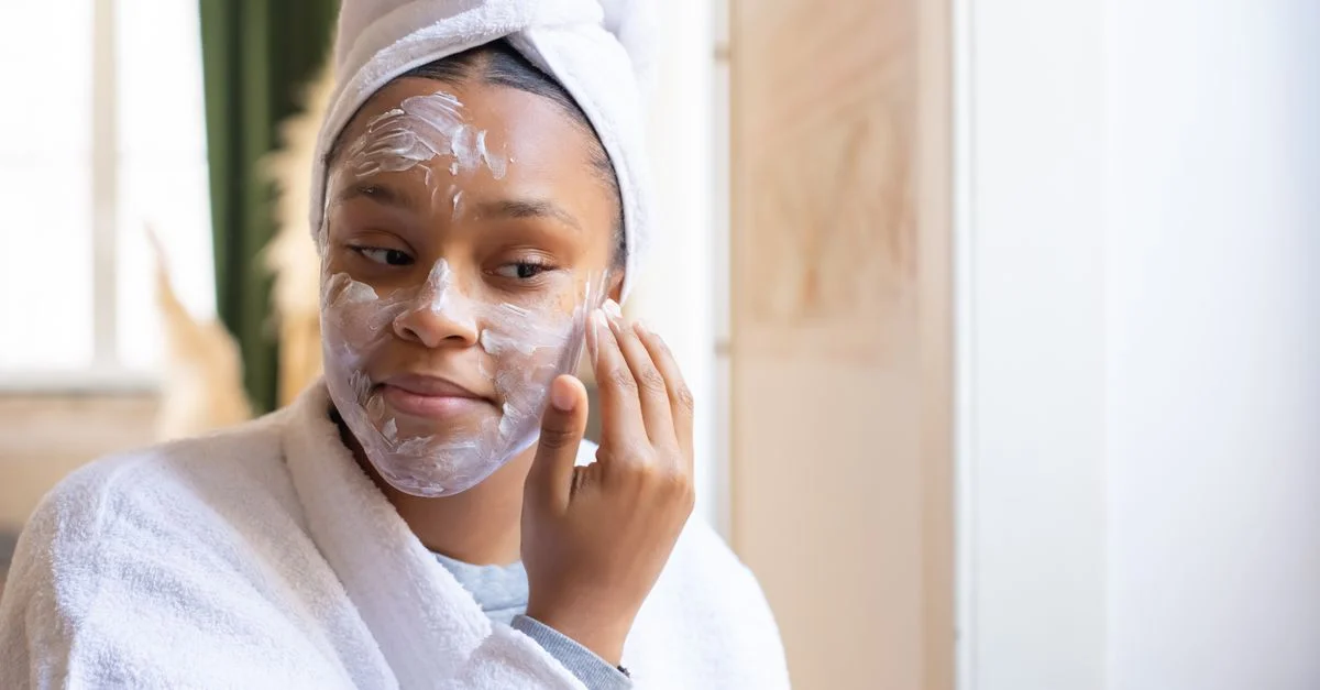 Une jeune femme en peignoir applique de la crème pour le visage tout en se relaxant à l'intérieur. Routine de soins de la peau matinale.