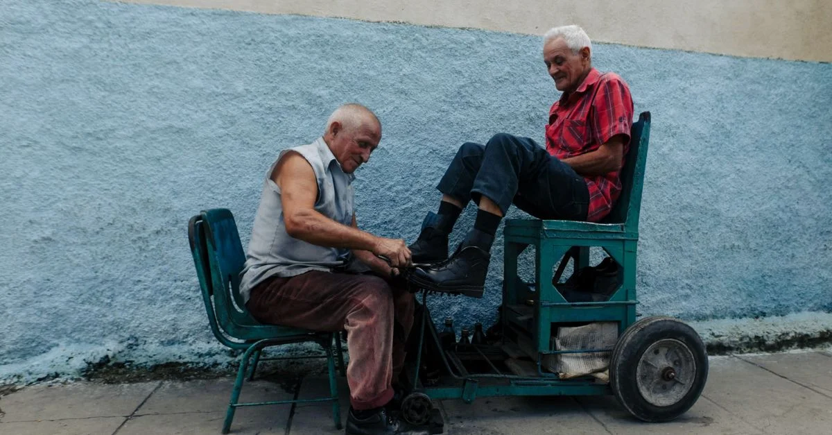 Deux hommes âgés interagissant en extérieur, l'un nettoyant les chaussures en cuir de l'autre sur un siège rustique.