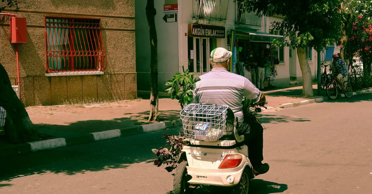 Un homme âgé roule sur un scooter de mobilité dans une rue ensoleillée avec des plantes dans le panier arrière.