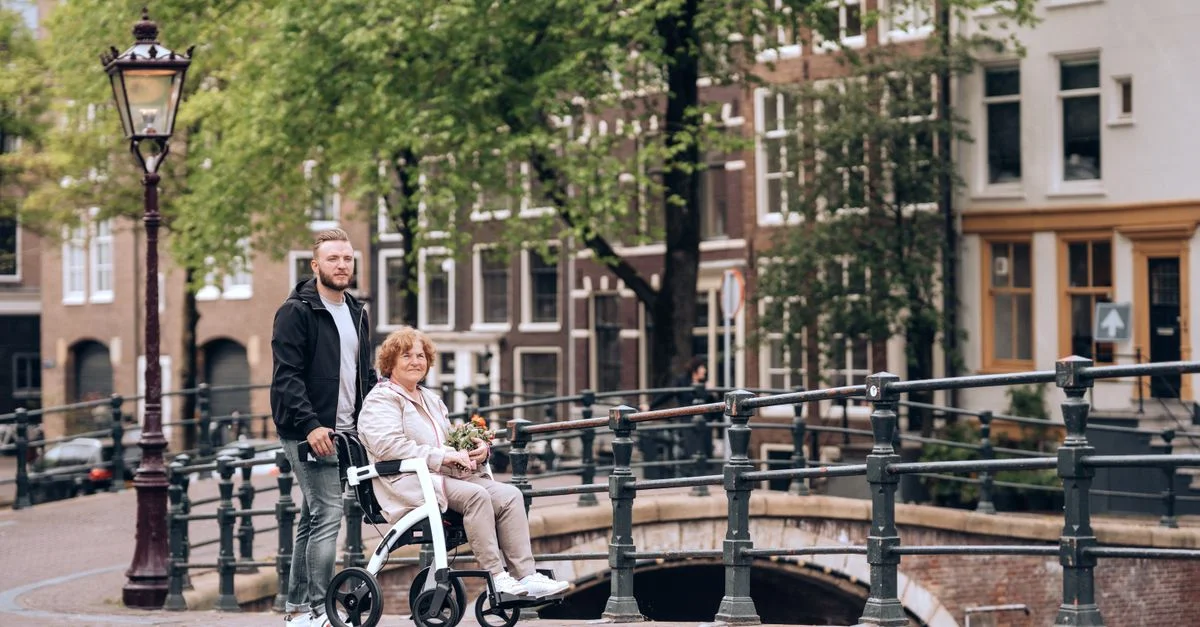 Femme âgée utilisant un déambulateur sur un pont canal à Amsterdam avec un jeune homme l'assistant. Scène urbaine.