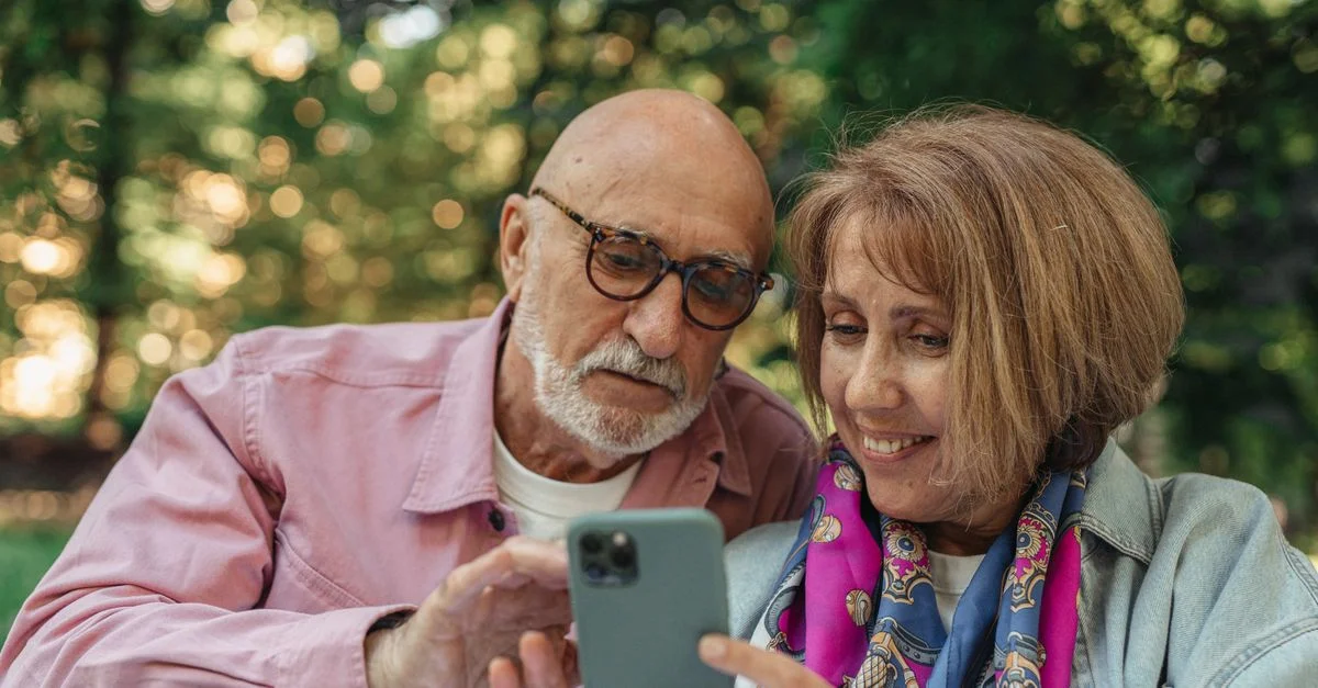 Couple de personnes âgées heureux utilisant un smartphone dans un parc, profitant du plein air ensoleillé.