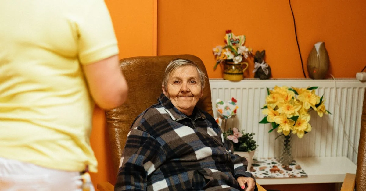 Une scène chaleureuse d'une femme senior assise dans un fauteuil dans une maison de retraite avec un soignant à proximité.