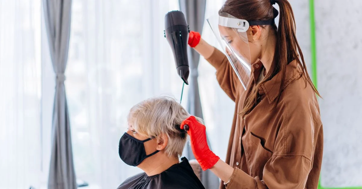 Une coiffeuse portant une visière et des gants sèche les cheveux d'un client dans un salon.