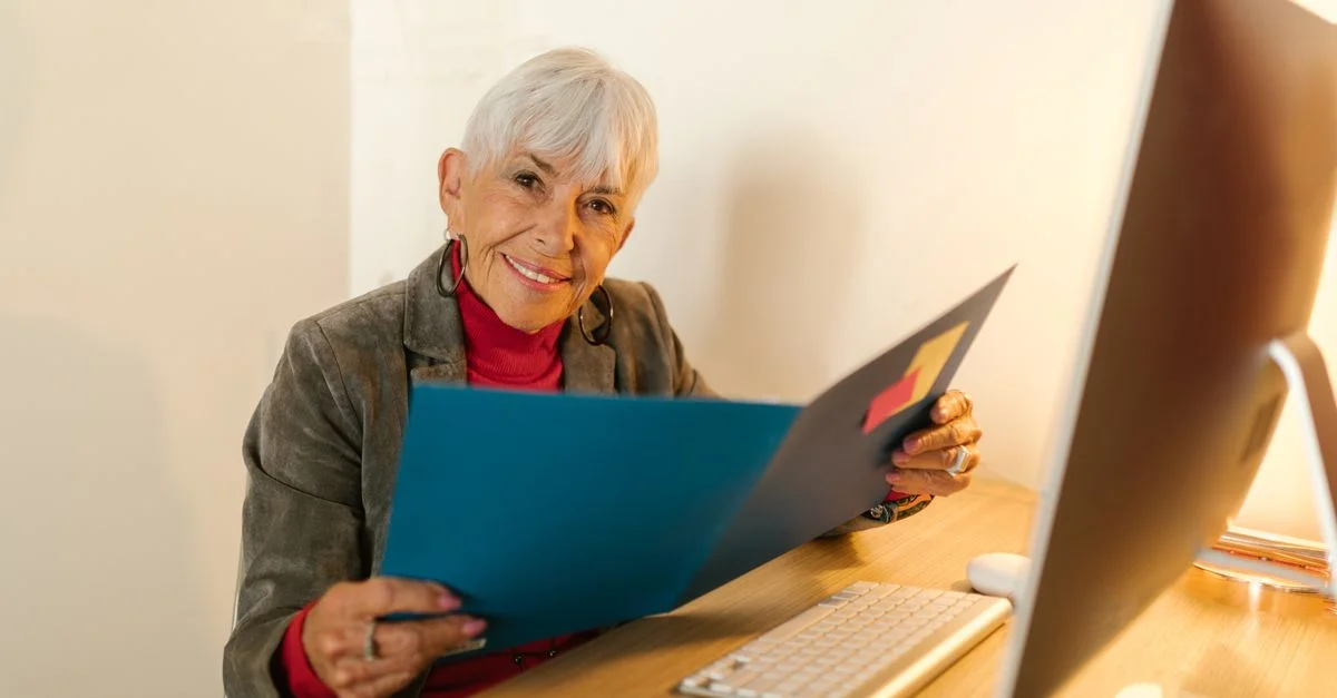 Femme âgée en tenue professionnelle assise à un bureau, tenant un dossier et souriant chaleureusement dans un cadre de bureau.