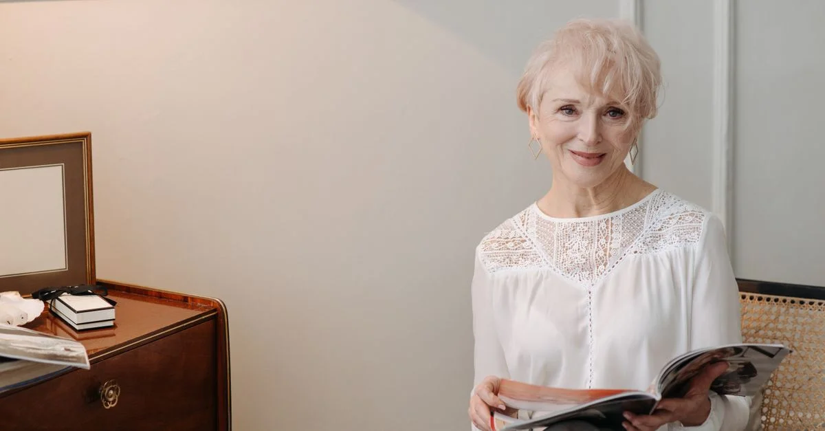 Une femme senior élégante assise à l'intérieur, profitant d'un magazine avec un sourire chaleureux.