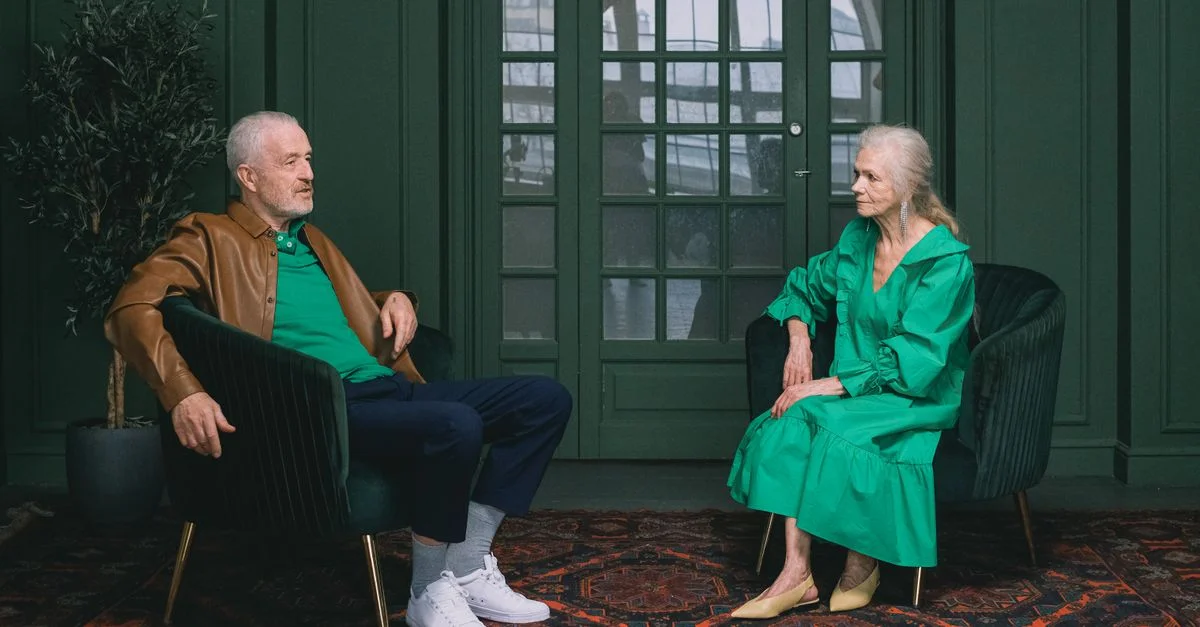 Homme et femme âgés dans une pièce aux thèmes verts engagés dans une conversation, assis sur des fauteuils.