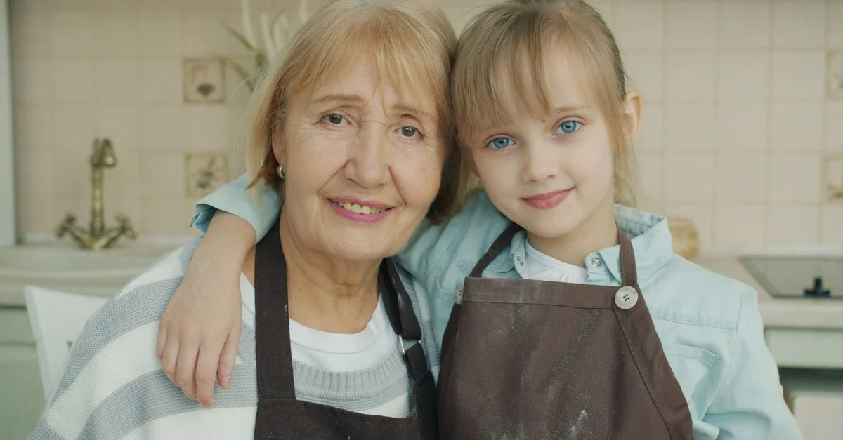 Une grand-mère et sa petite-fille en tablier, partageant un moment de complicité dans la cuisine.