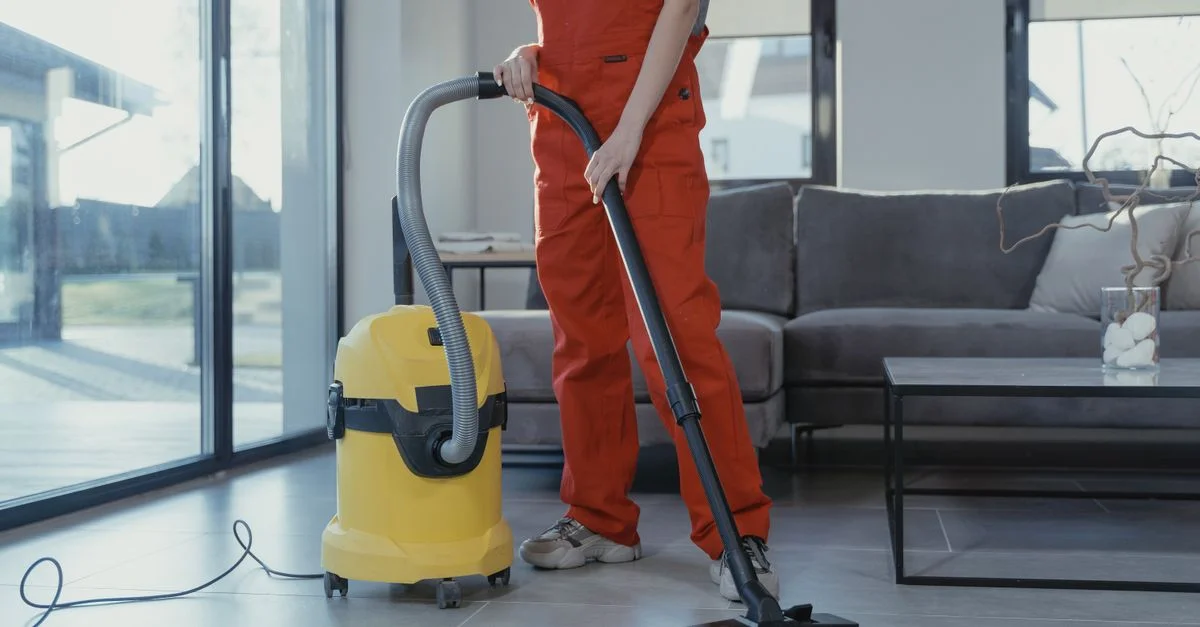 Un nettoyeur professionnel en combinaison rouge aspirant un salon moderne avec un aspirateur jaune.