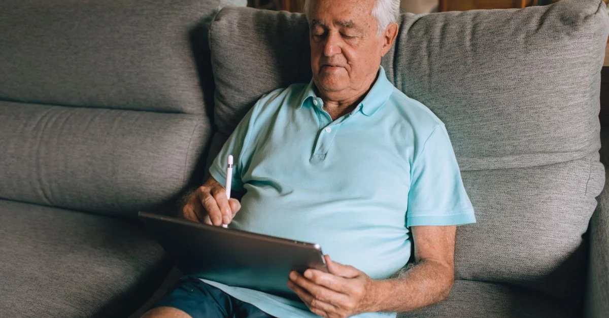 Homme âgé en polo bleu utilisant une tablette tout en se relaxant sur un canapé confortable.