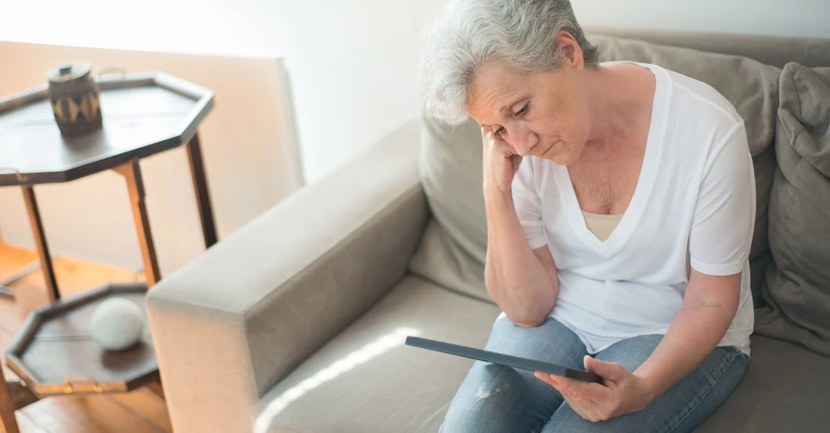 Une femme senior est assise pensivement sur un canapé dans un salon lumineux, tenant une tablette.