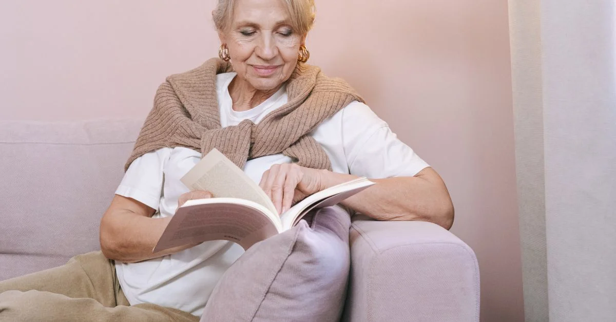 Femme âgée appréciant un livre à l'intérieur, assise confortablement sur un canapé de couleur pastel.