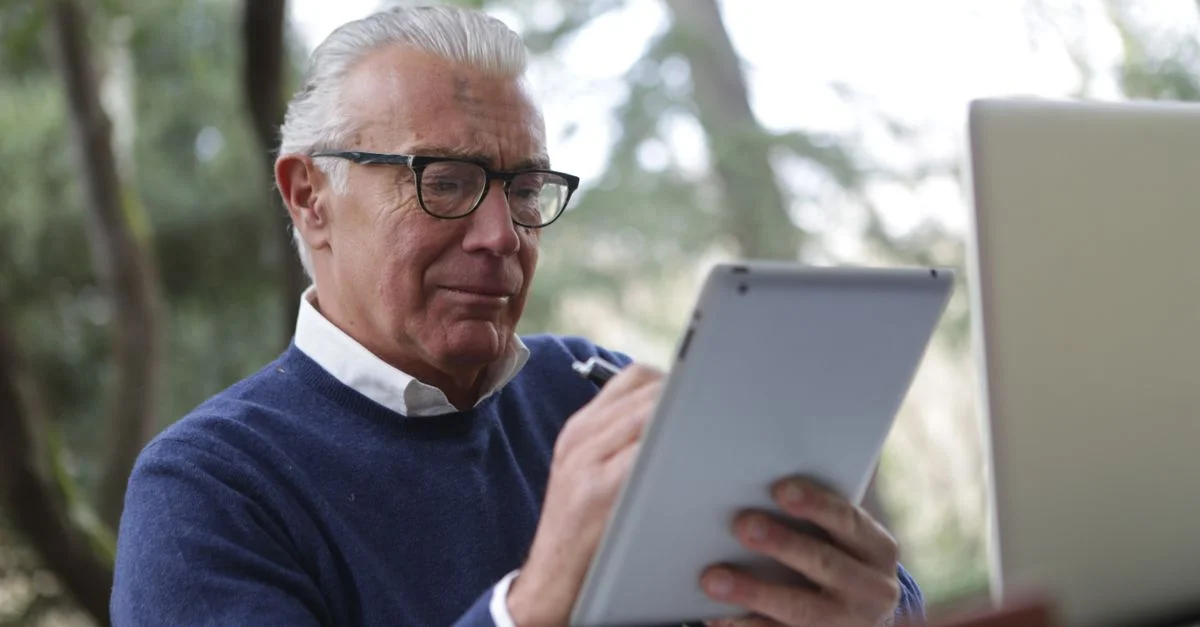 Un homme âgé utilisant une tablette et un ordinateur portable en plein air, indiquant un style de vie moderne des seniors.