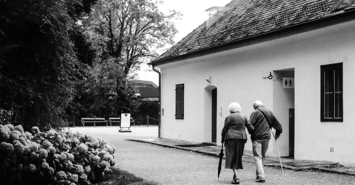 Une photographie en noir et blanc d'un couple de personnes âgées marchant sur un chemin devant un bâtiment blanc.