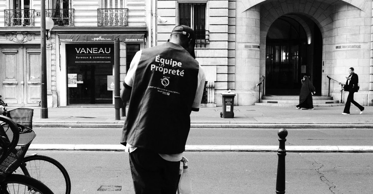 Une photo en noir et blanc d'un travailleur urbain nettoyant une rue animée, capturant l'activité piétonnière variée.