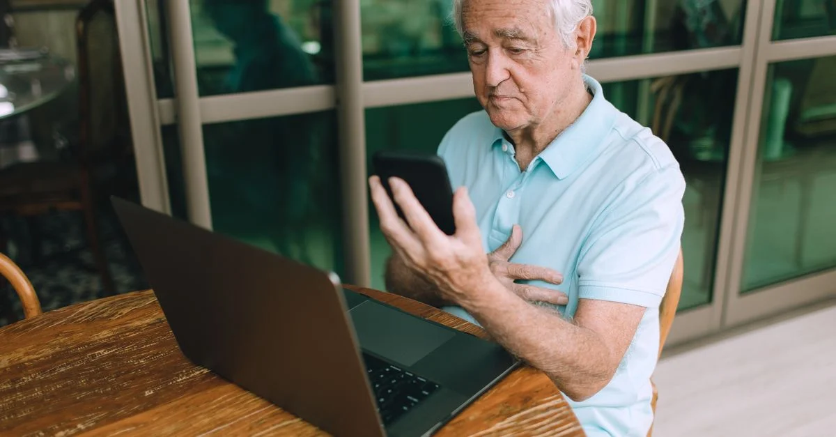 Homme âgé assis à une table utilisant un smartphone et un ordinateur portable, illustrant l'utilisation de la technologie.