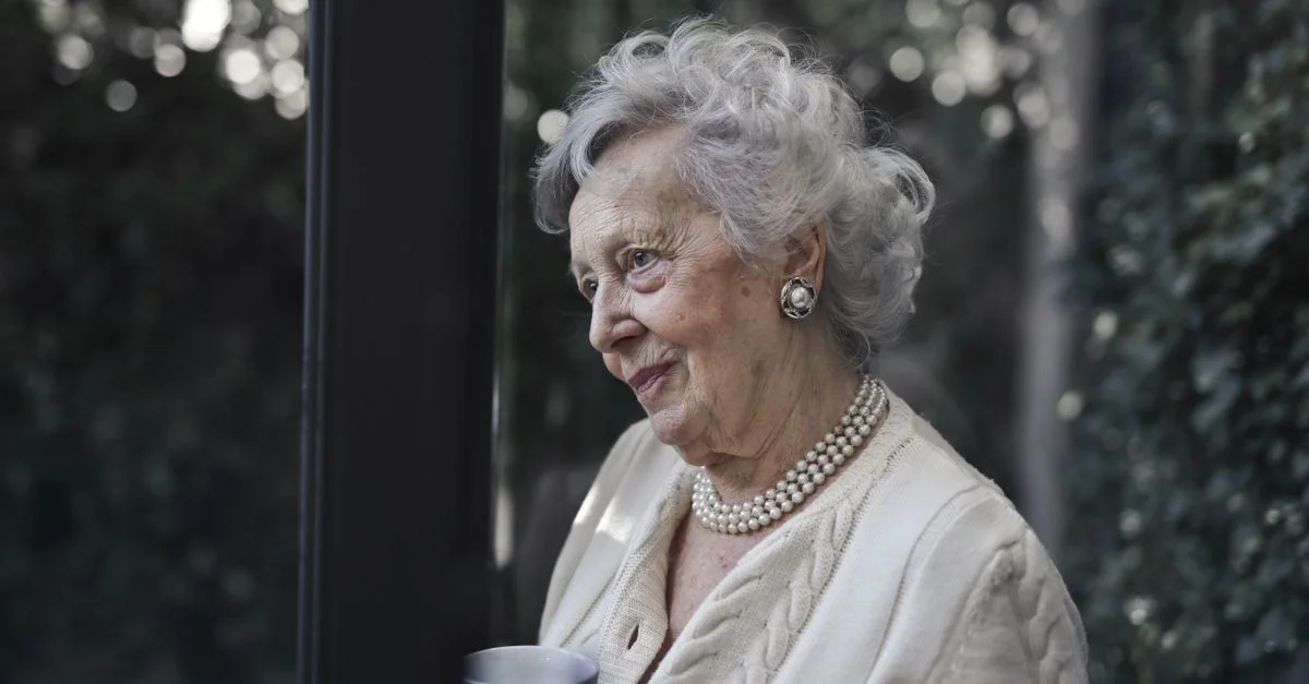 Femme senior aux cheveux blancs et aux perles profitant d'un moment tranquille à l'intérieur.