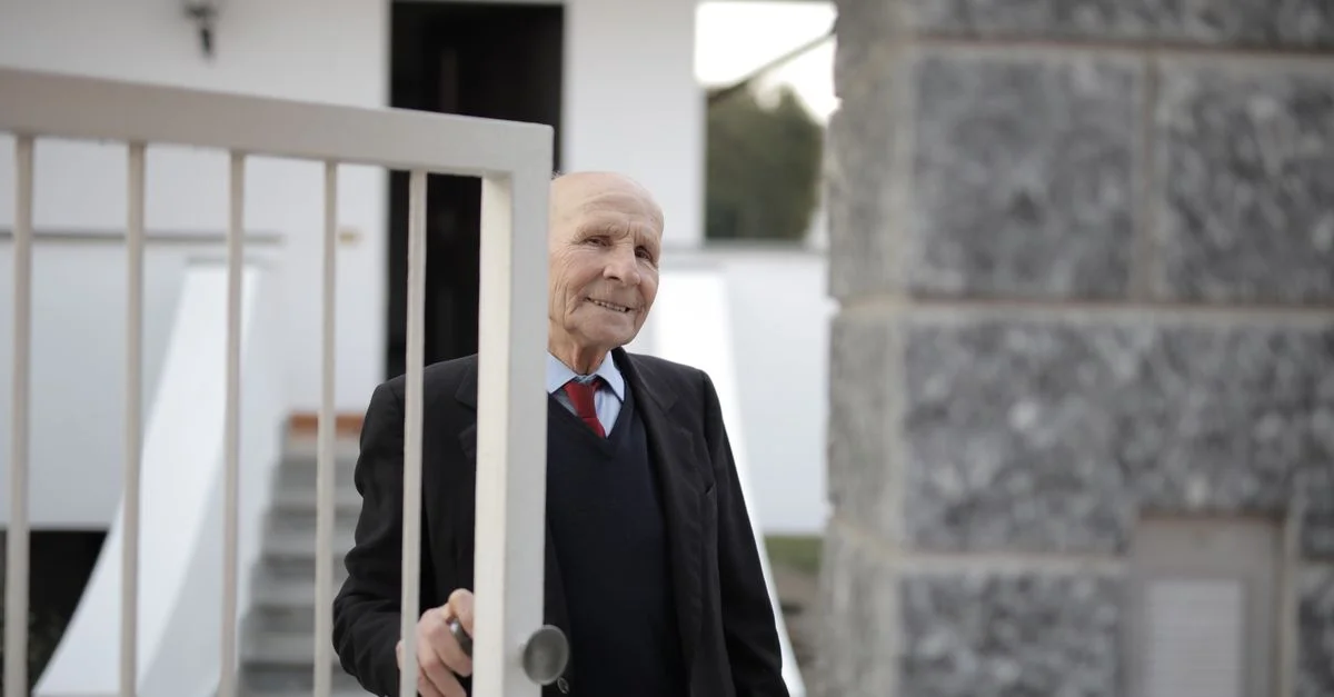Un homme âgé en costume ouvre joyeusement le portail de sa maison, profitant de la lumière du jour.