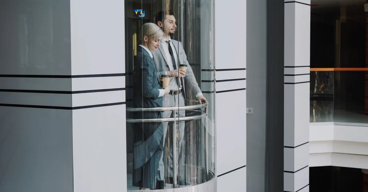 Deux professionnels en affaires en costume se tenant à l'intérieur d'un ascenseur en verre moderne.