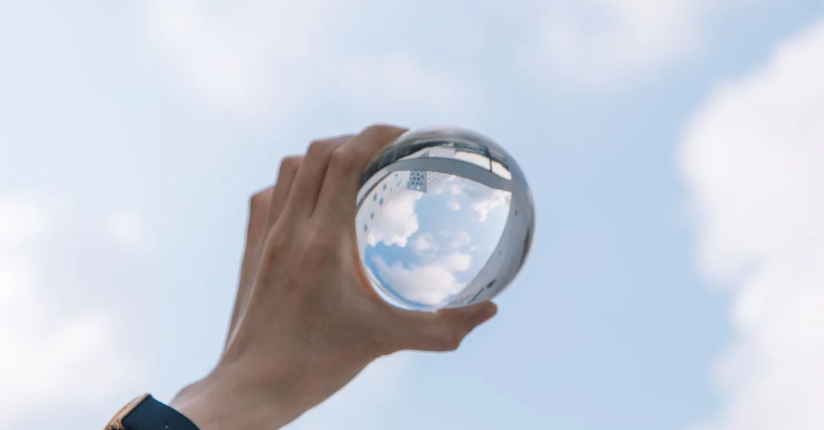Une main tient une boule en cristal reflétant le ciel et les nuages, créant une image inversée intrigante.