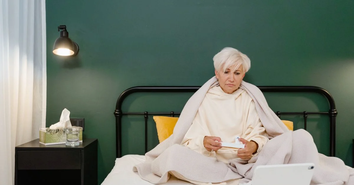 Femme senior au lit utilisant une tablette pour la télémédecine pendant la maladie, enveloppée dans une couverture.