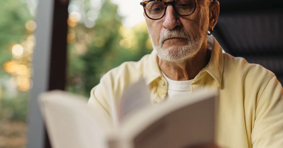 Homme âgé avec des lunettes, concentré sur la lecture d'un livre à l'extérieur.