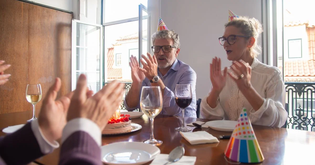 Un couple senior célèbre un anniversaire en intérieur avec des amis, portant des chapeaux de fête et profitant du gâteau.