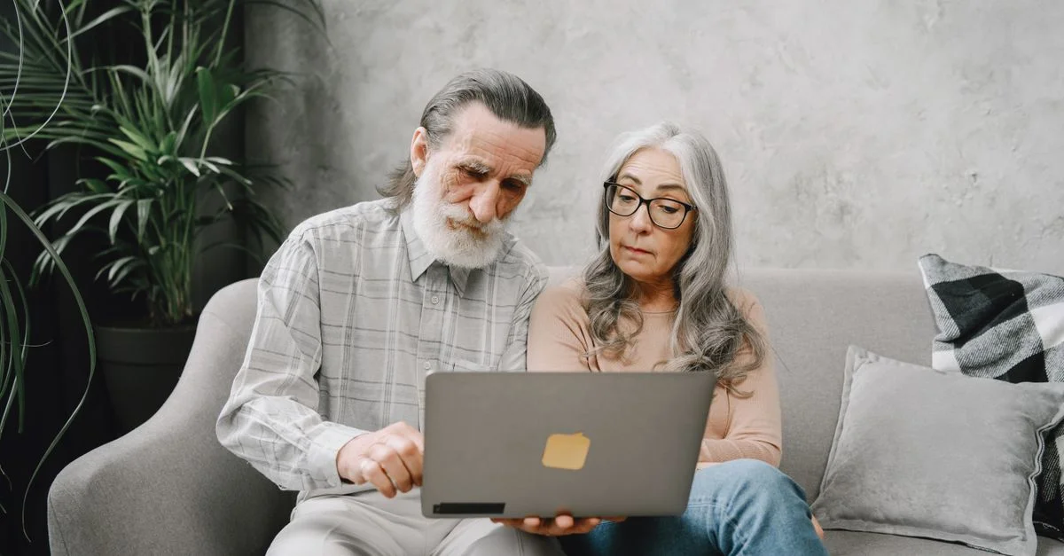 Couple de seniors assis sur un canapé utilisant un ordinateur portable à l'intérieur, adoptant la technologie ensemble.