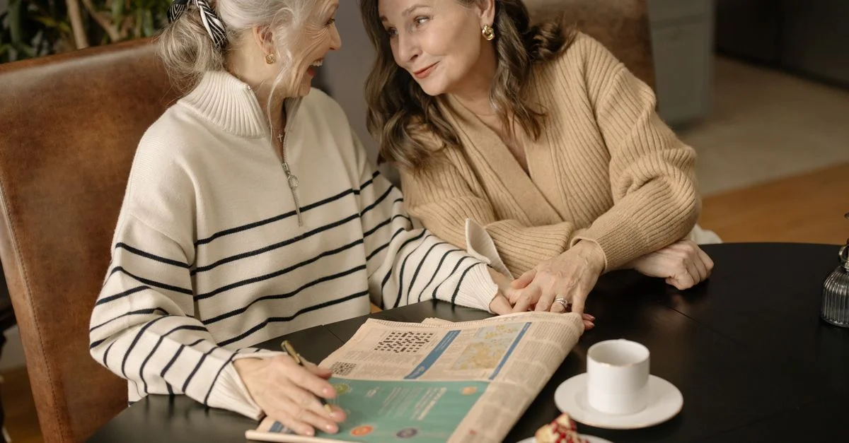 Deux femmes âgées partagent un moment autour d'un café et d'un mots croisés à l'intérieur.