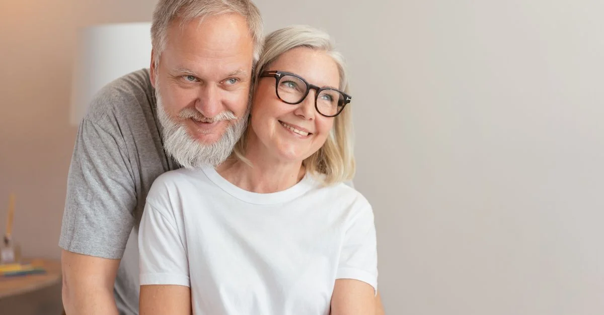 Couple âgé souriant partageant une étreinte affectueuse à l'intérieur, tous deux portant des lunettes.