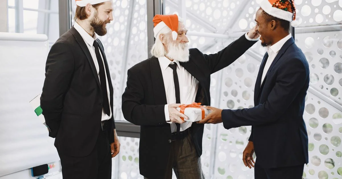 Trois collègues en costume échangent des cadeaux de Noël dans un bureau festif.