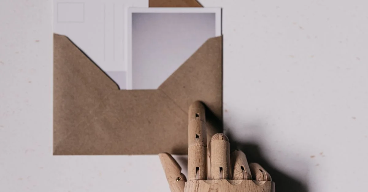 Une main de mannequin en bois tient une enveloppe brune contenant des cartes postales blanches.