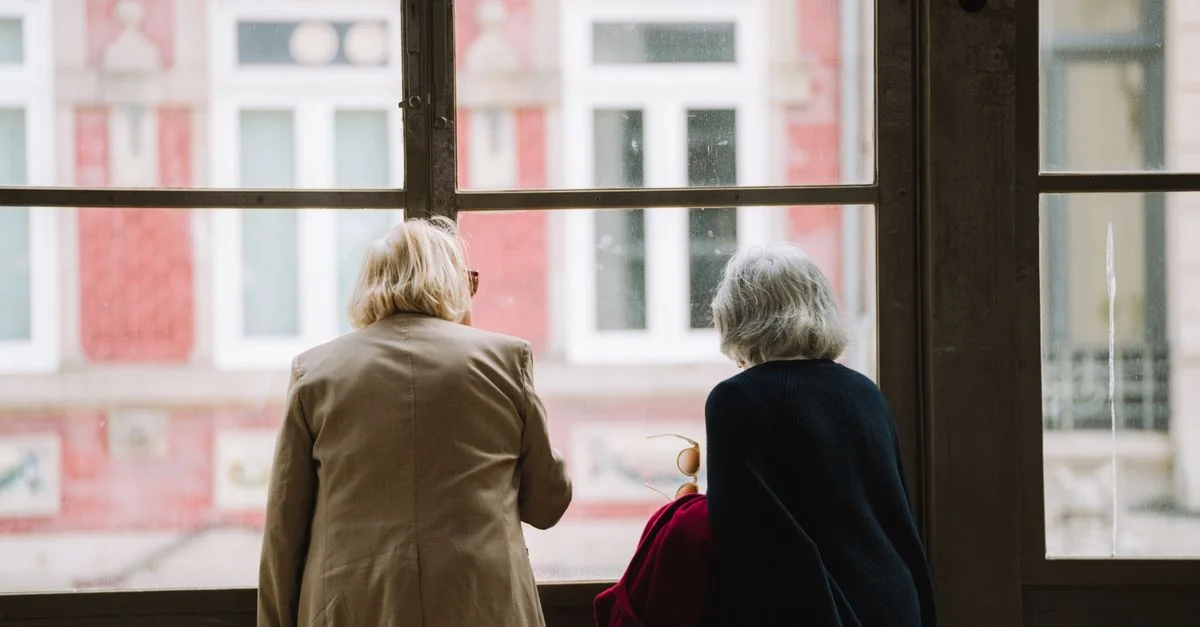 Deux femmes âgées aux cheveux gris regardent par la fenêtre une scène de ville, capturant un moment serein.