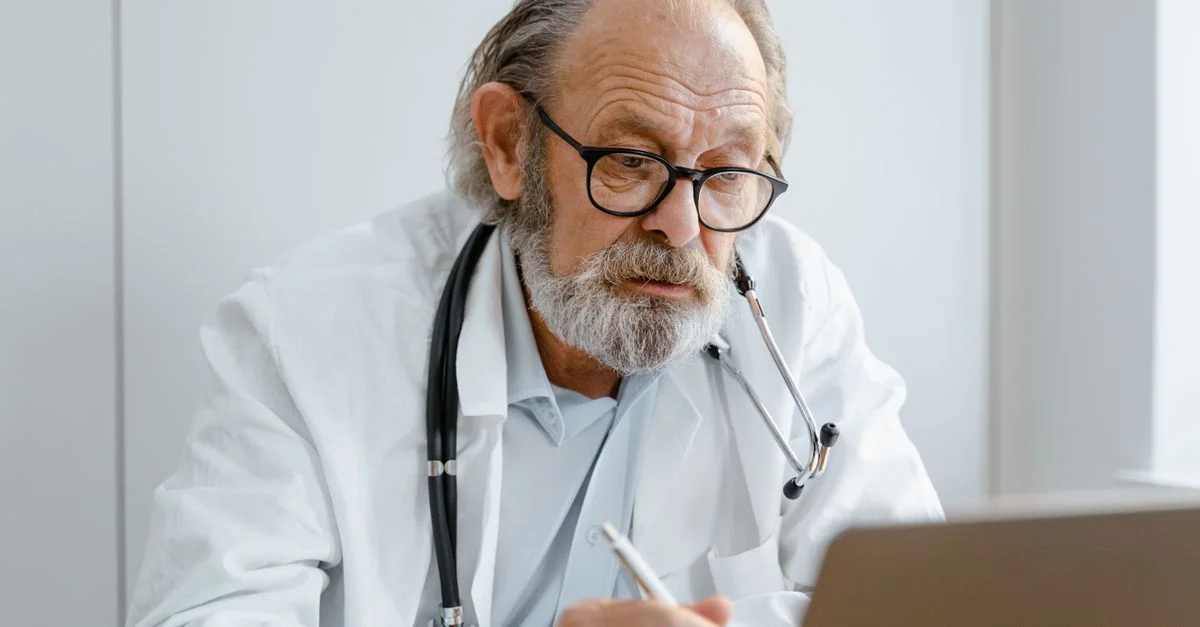 Médecin âgé avec des lunettes et un stéthoscope travaillant sur un ordinateur portable dans un cabinet médical.