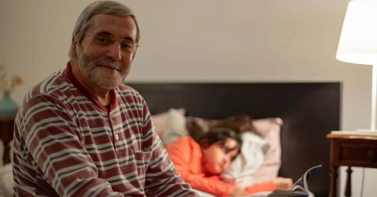 Homme senior lisant dans une chambre confortable pendant qu'un jeune se repose à proximité, capturant un moment familial chaleureux.