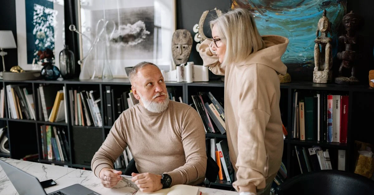 Un couple âgé discutant de quelque chose autour d'une table en marbre avec des livres et un ordinateur portable, entouré d'art et de sculptures.