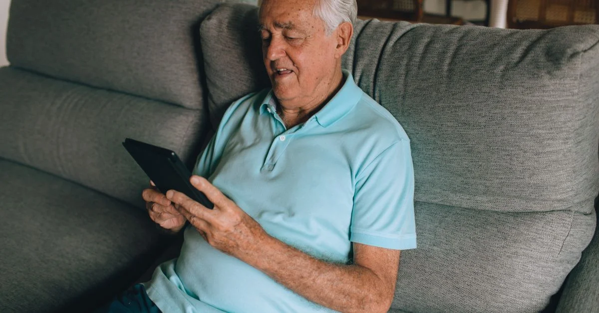Un homme senior souriant avec des cheveux gris utilisant une tablette sur un canapé à l'intérieur.