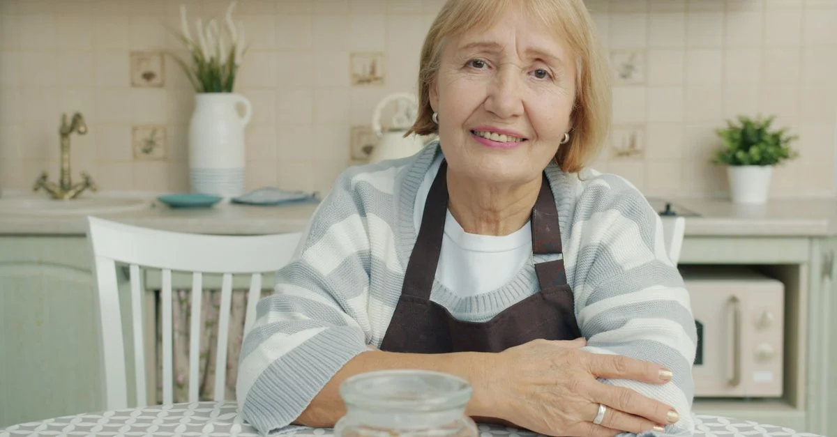 Une femme âgée en tablier, assise dans une cuisine chaleureuse, sourit.