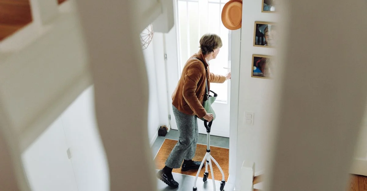 Une femme âgée utilise un déambulateur à l'intérieur près de la porte, se préparant à quitter la maison.