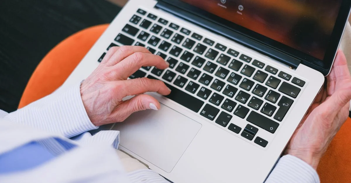 Un senior utilisant un clavier d'ordinateur portable, portant une chemise rayée, assis à l'intérieur.