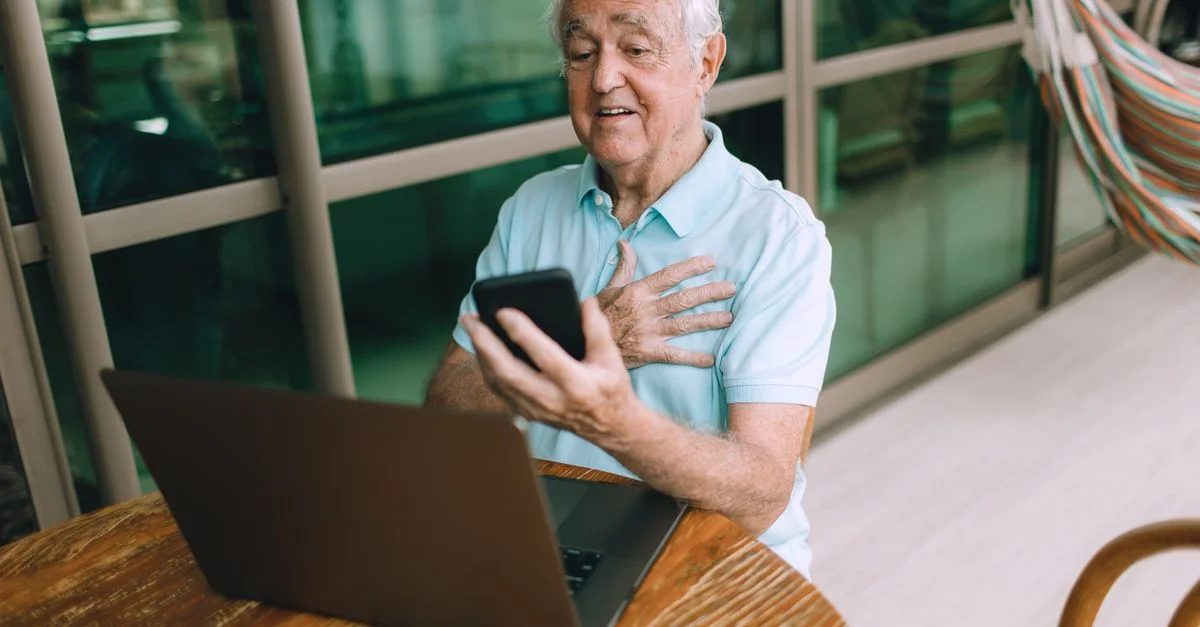Homme âgé utilisant un smartphone et un ordinateur portable pour un appel vidéo à la maison, engagé avec la technologie.