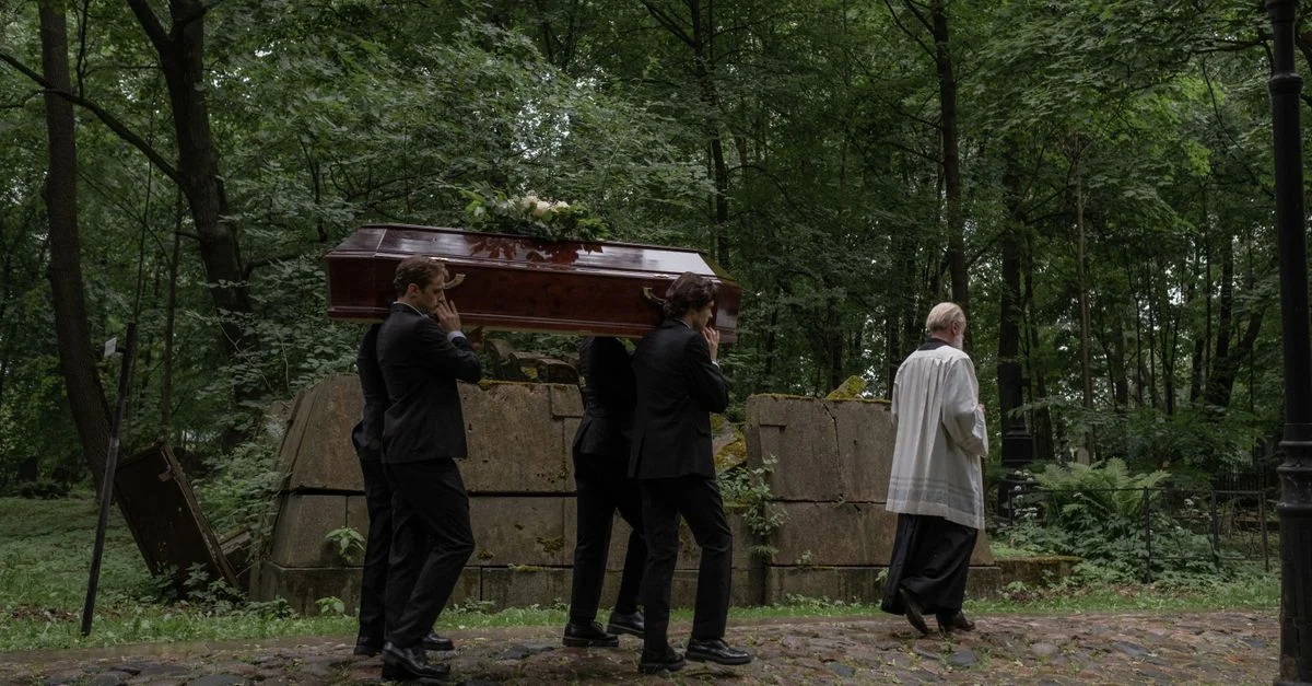 Des hommes en costume noir portent un cercueil en bois lors d'un cortège funèbre dans un cimetière forestier.