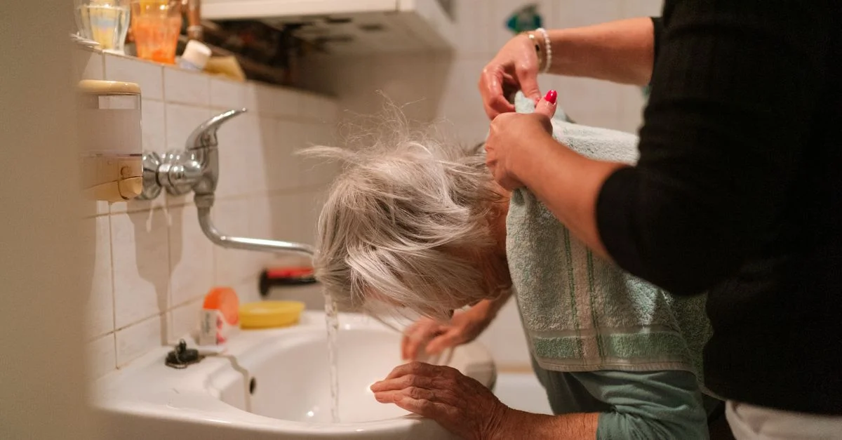 Femme âgée aux cheveux gris recevant de l'aide d'un soignant dans une salle de bains.