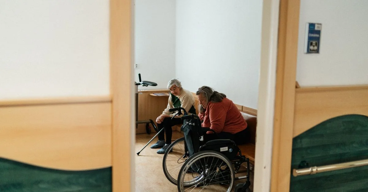 Deux femmes seniors conversent à l'intérieur, l'une dans un fauteuil roulant, symbolisant la convivialité dans un cadre de maison de retraite.